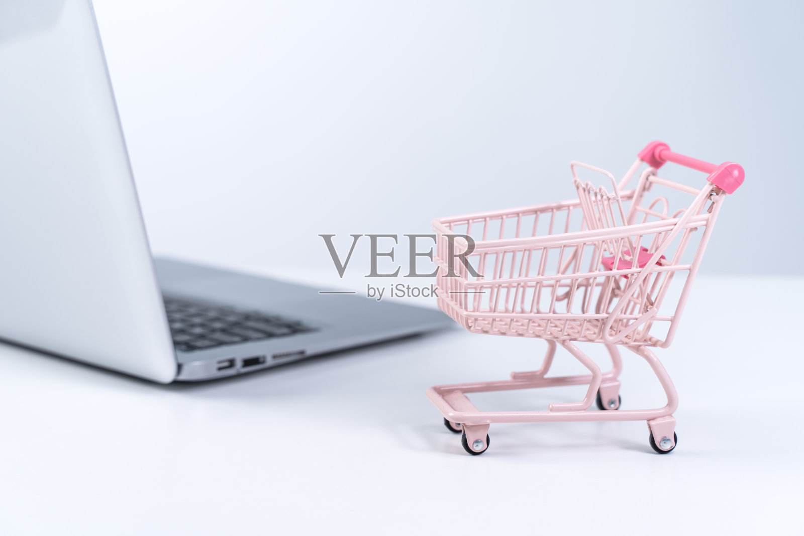 在线购物。迷你空粉色购物车购物车上方的笔记本电脑在白色的桌子背景，购买在家的概念。照片摄影图片