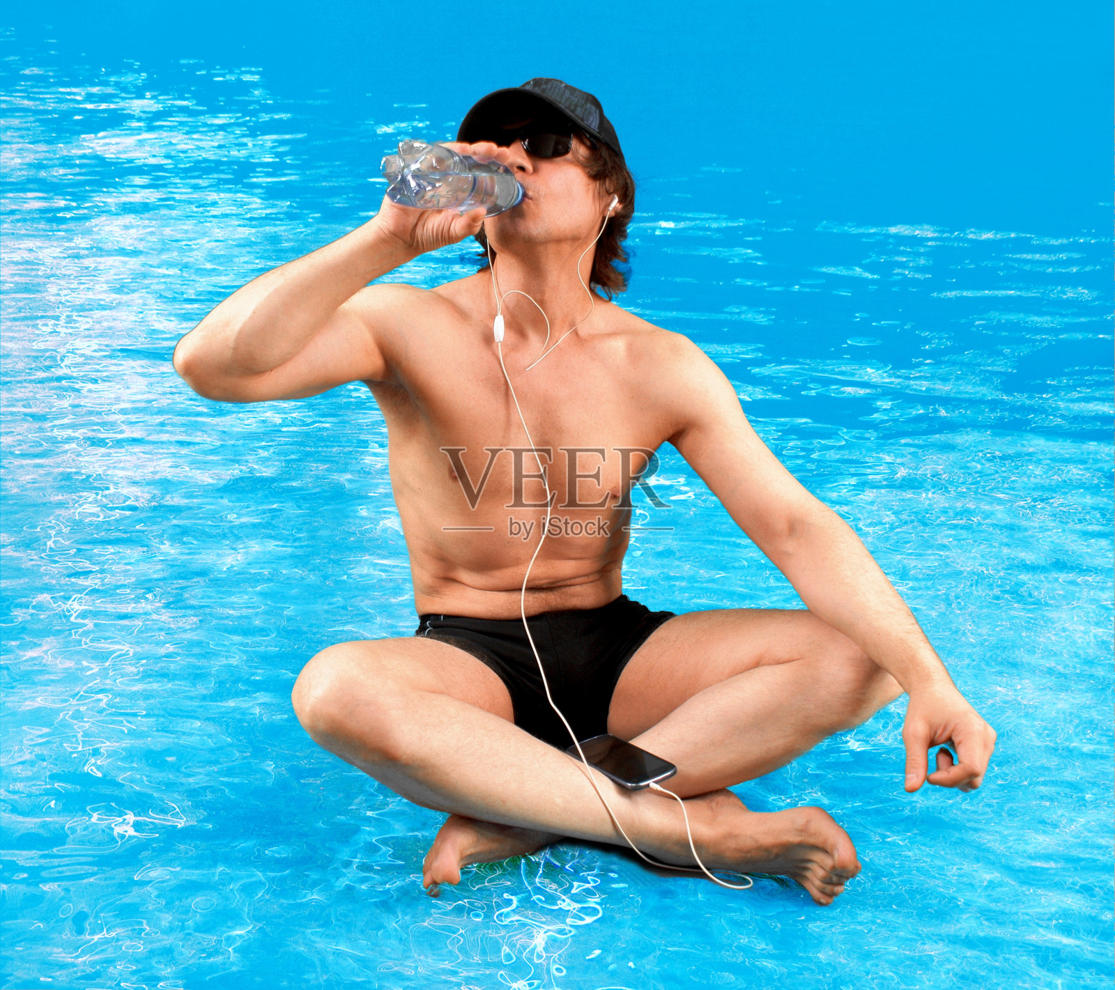 有魅力的运动员盘腿坐在水中，晒日光浴，用智能手机听音乐，用瓶子喝水。照片摄影图片