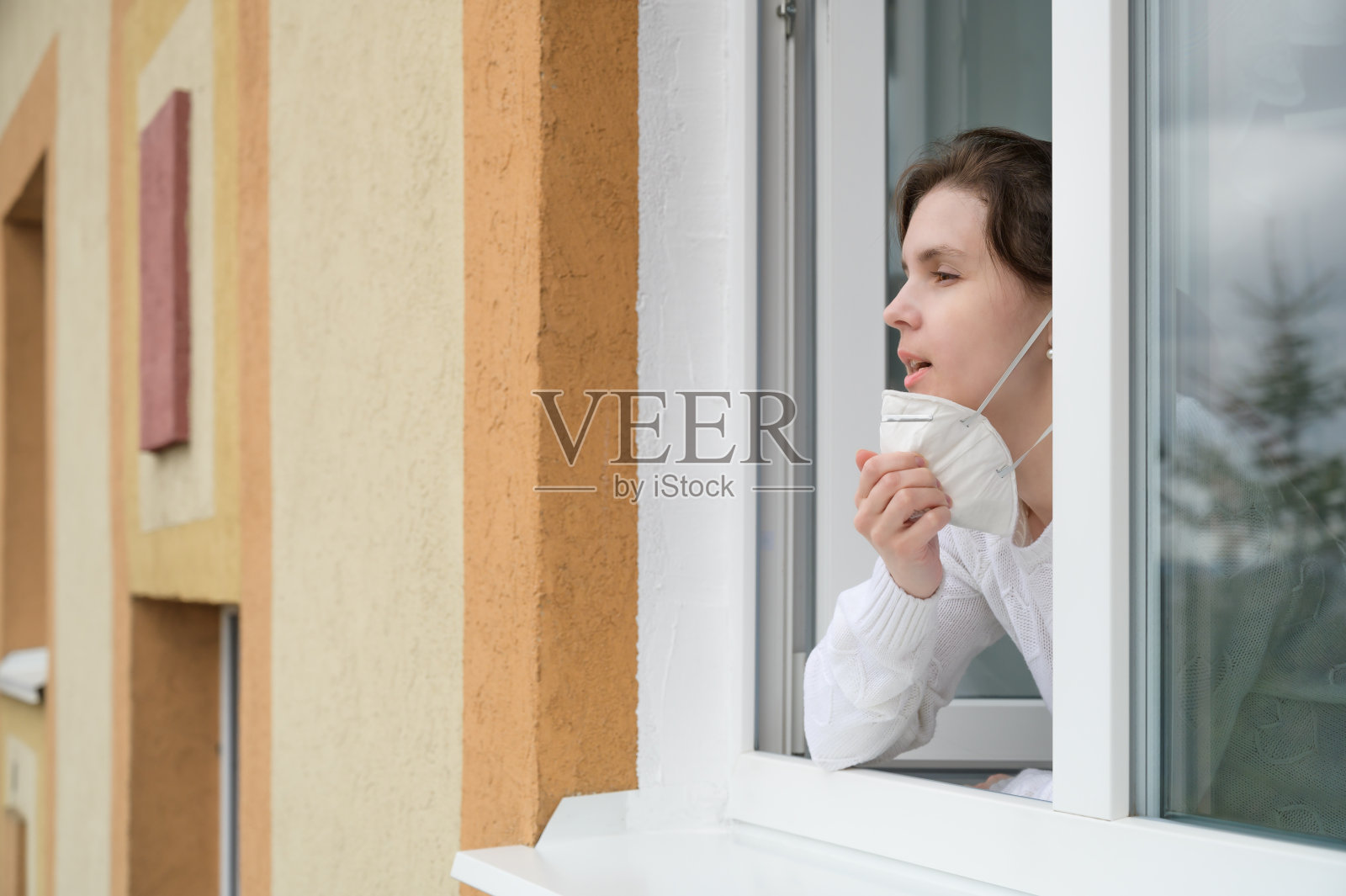 一名年轻的白人女子看着公寓窗外，摘下了她的呼吸器。照片摄影图片