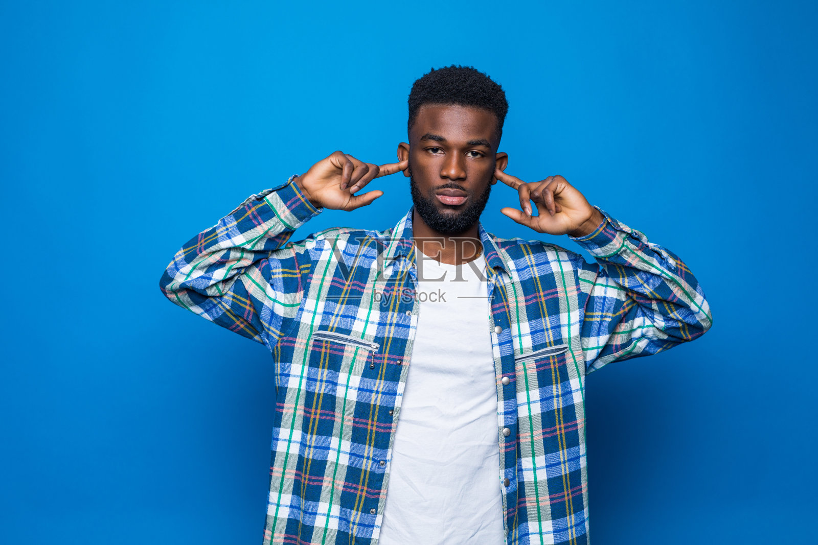 非裔美国人，胡须遮住耳朵，不理会恼人的噪音，用耳塞堵住耳朵以避免听到蓝色背景下隔离的声音。嘈杂的音乐是个问题。照片摄影图片