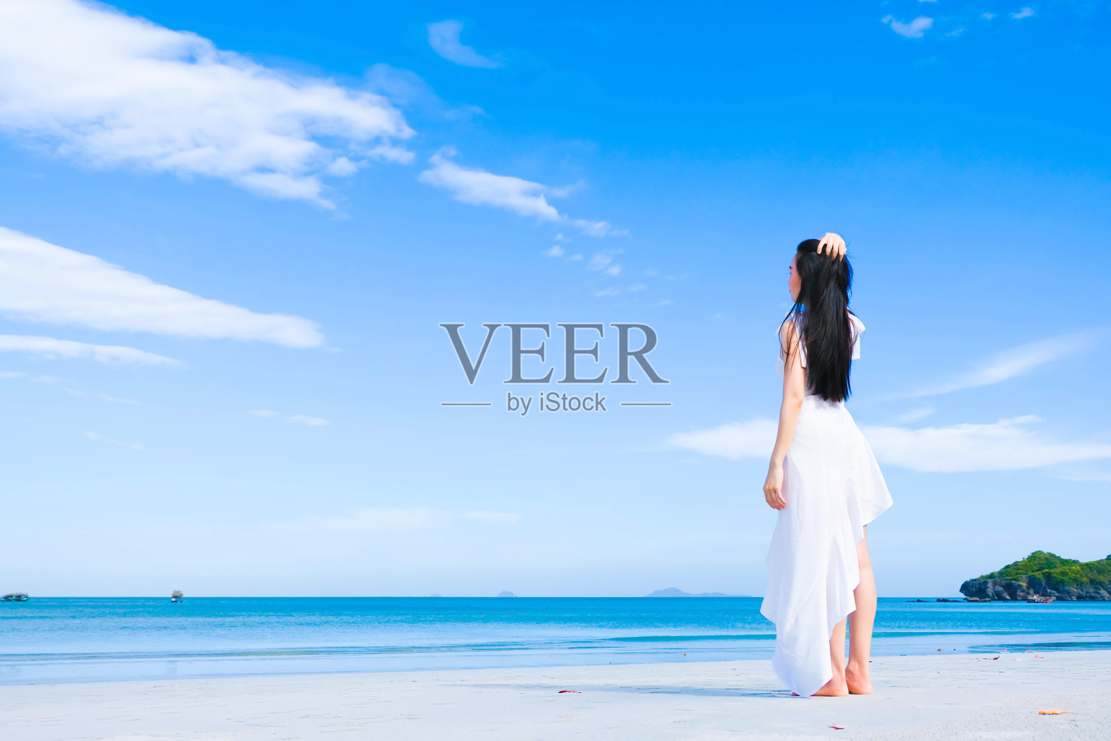 亚洲女性，长长的黑发，穿着白色的连衣裙和帽子站在海滩附近编织的袋子和太阳镜，背对着大海与拷贝空间蓝天，暑假和度假的概念照片摄影图片