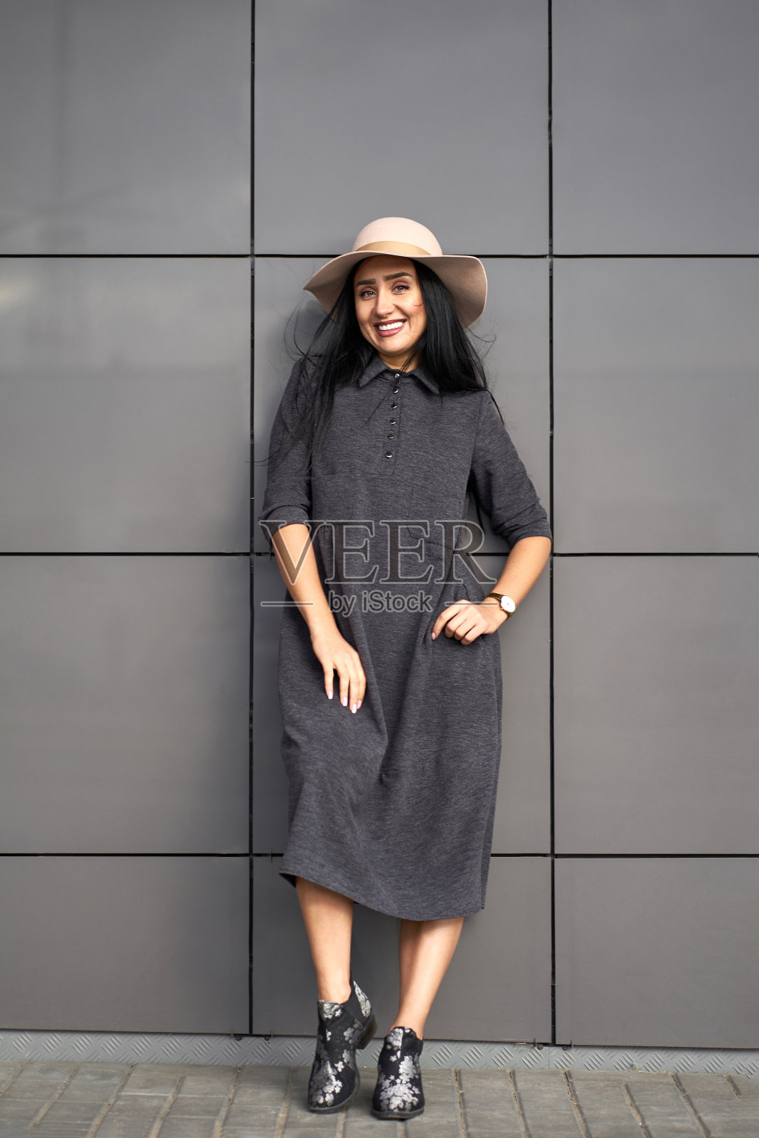 新时尚系列。幸福美丽的黑发女孩穿着时尚的灰色连衣裙，手里拿着时尚的帽子。模特在时尚的服装站在灰色的墙壁背景。户外肖像。照片摄影图片