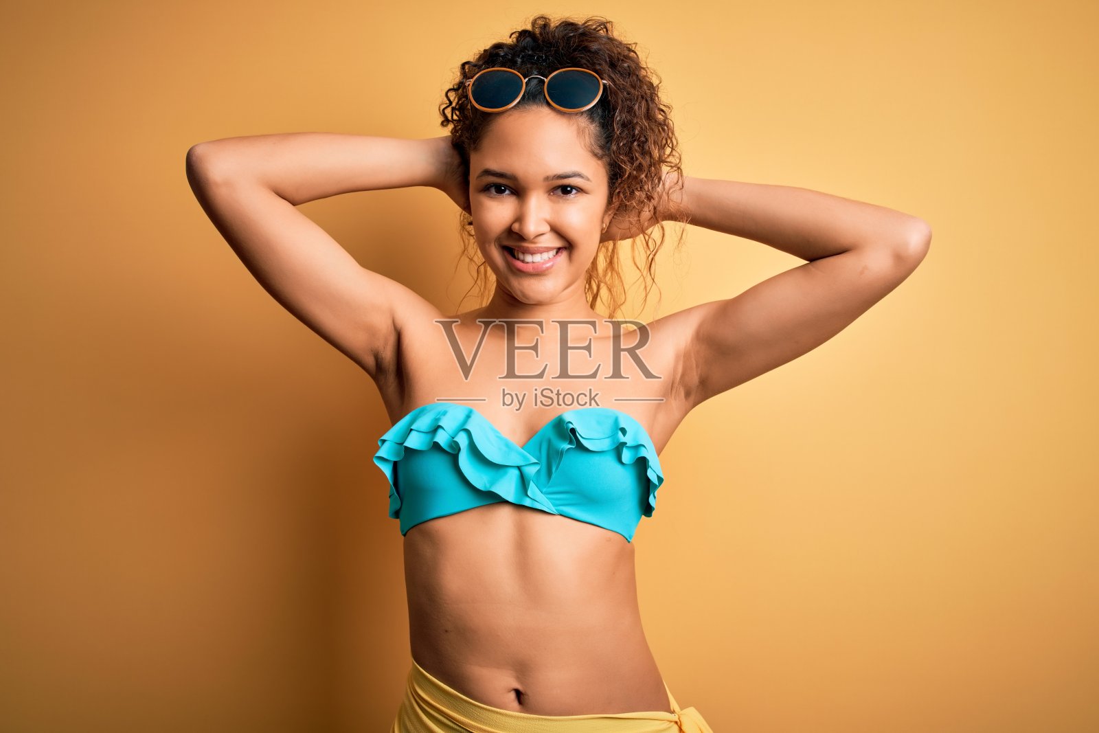 年轻美丽的美国女人在假期穿着比基尼在孤立的黄色背景放松和伸展，手臂和双手后的头和脖子微笑幸福照片摄影图片