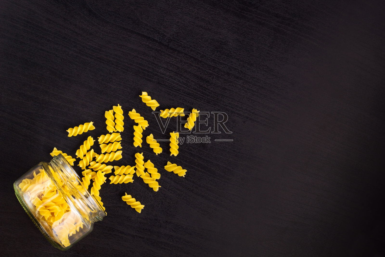 玻璃罐子里装满了干意面螺旋面，散落在黑色的背景上照片摄影图片