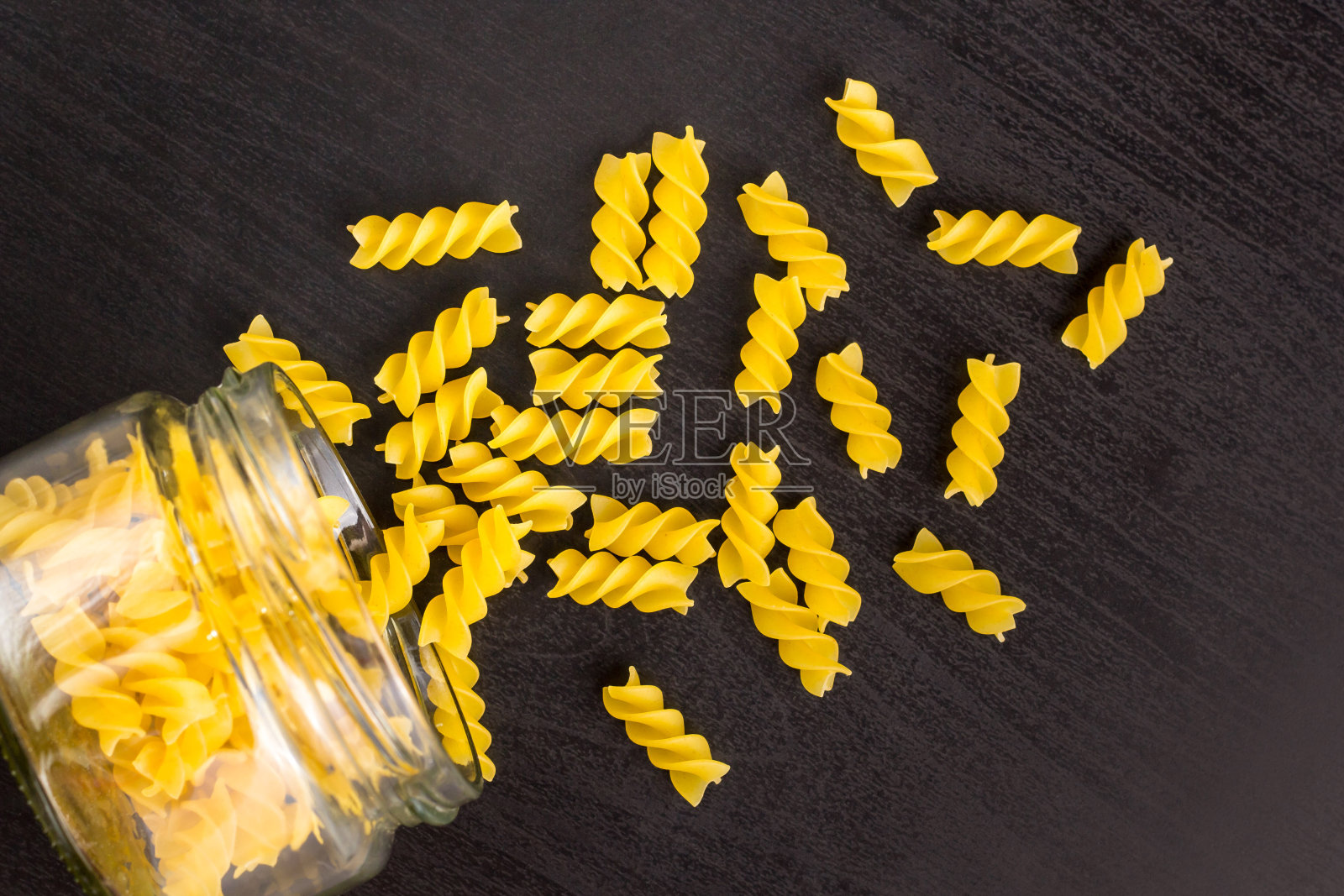 玻璃罐子里装满了干意面螺旋面，散落在黑色的背景上照片摄影图片