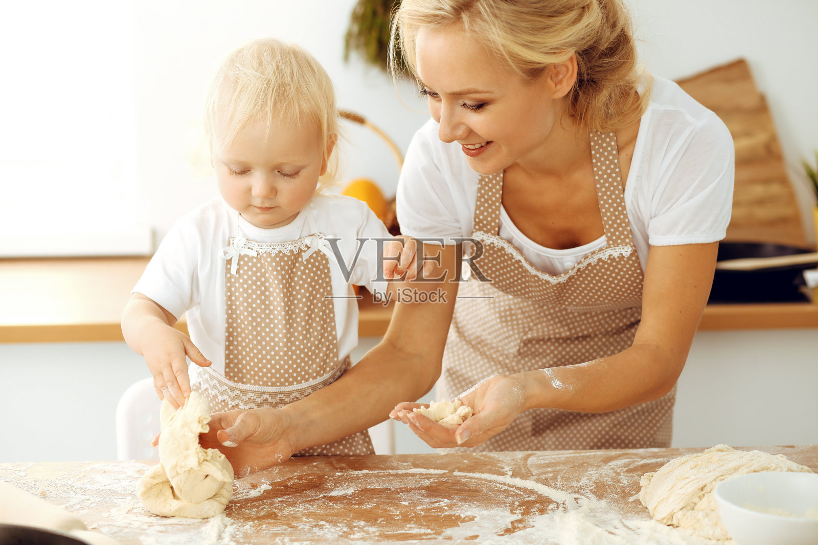 小女孩和她金发碧眼的妈妈穿着米色围裙，一边在厨房揉面，一边又玩又笑。自制的面包糕点，比萨饼或烤饼干。家庭乐趣和烹饪理念照片摄影图片