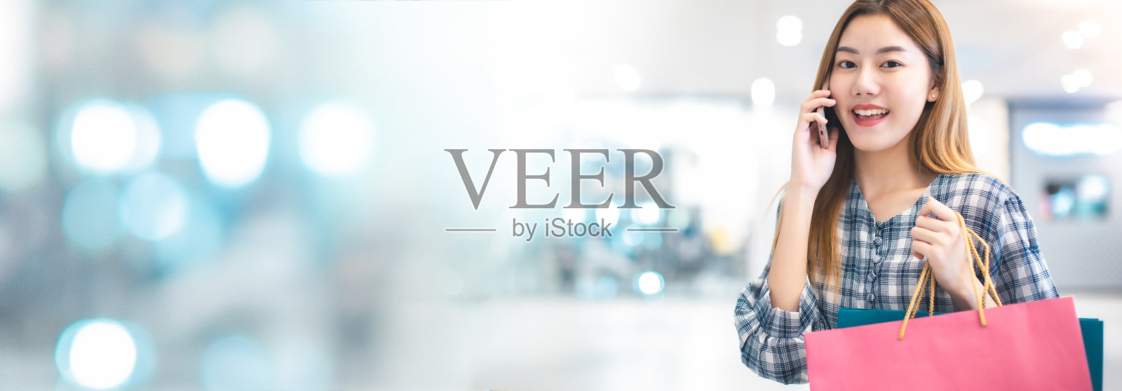 微笑年轻的亚洲女人与购物彩袋在商场背景。用智能手机在网上购物，站在商场大楼里微笑。生活方式概念，横幅图形照片摄影图片