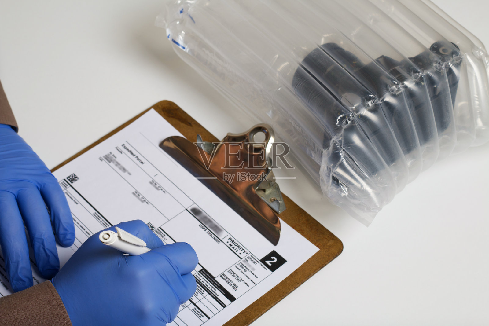 自我隔离期间的包裹递送。一名戴着橡胶手套的男子在送货单上签字。附近的一个气泡袋里装着一个摄像头。照片摄影图片