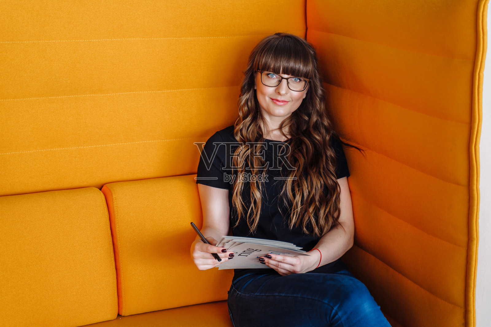 一个年轻的女孩坐在一个黄色的大沙发上。穿着便服——黑色t恤和蓝色牛仔裤，手里拿着教育卡片。在家庭之外工作的自由职业者。远程教育学生照片摄影图片