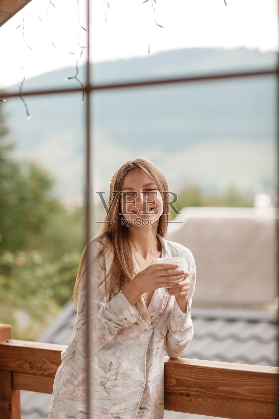 年轻女子在阳台上拿着一杯咖啡或茶在早上。夏天，她在旅馆的房间里观赏大自然。女孩穿着时髦的睡衣。放松的时间。照片摄影图片