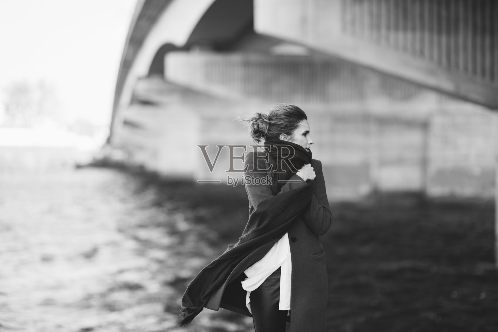 穿着大衣和围巾的美丽孤独的女人在河边的桥下随风飘动。孤独的概念。黑白艺术照片。软的焦点。照片摄影图片