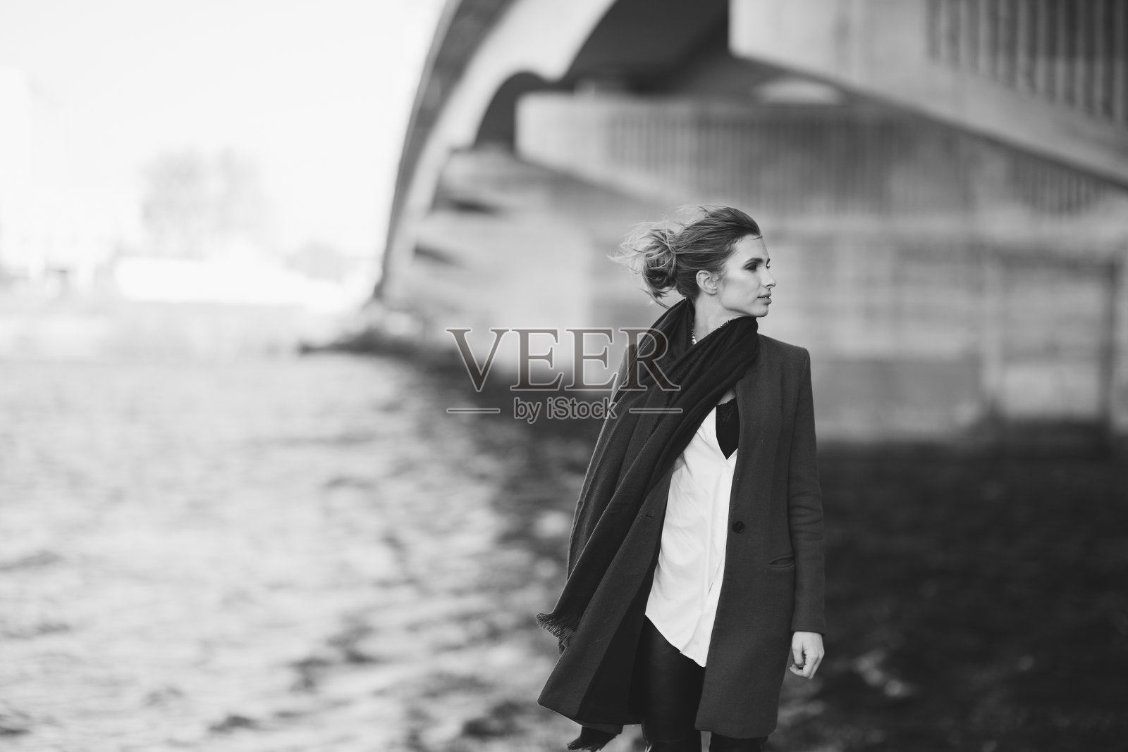 穿着大衣和围巾的美丽孤独的女人在河边的桥下随风飘动。孤独的概念。黑白艺术照片。软的焦点。照片摄影图片