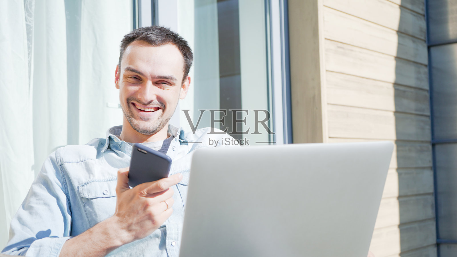微笑英俊的男子在电话与他的家人交谈。微笑的休闲商人有一个日光浴。在家中阳台上使用笔记本电脑自我隔离。照片摄影图片
