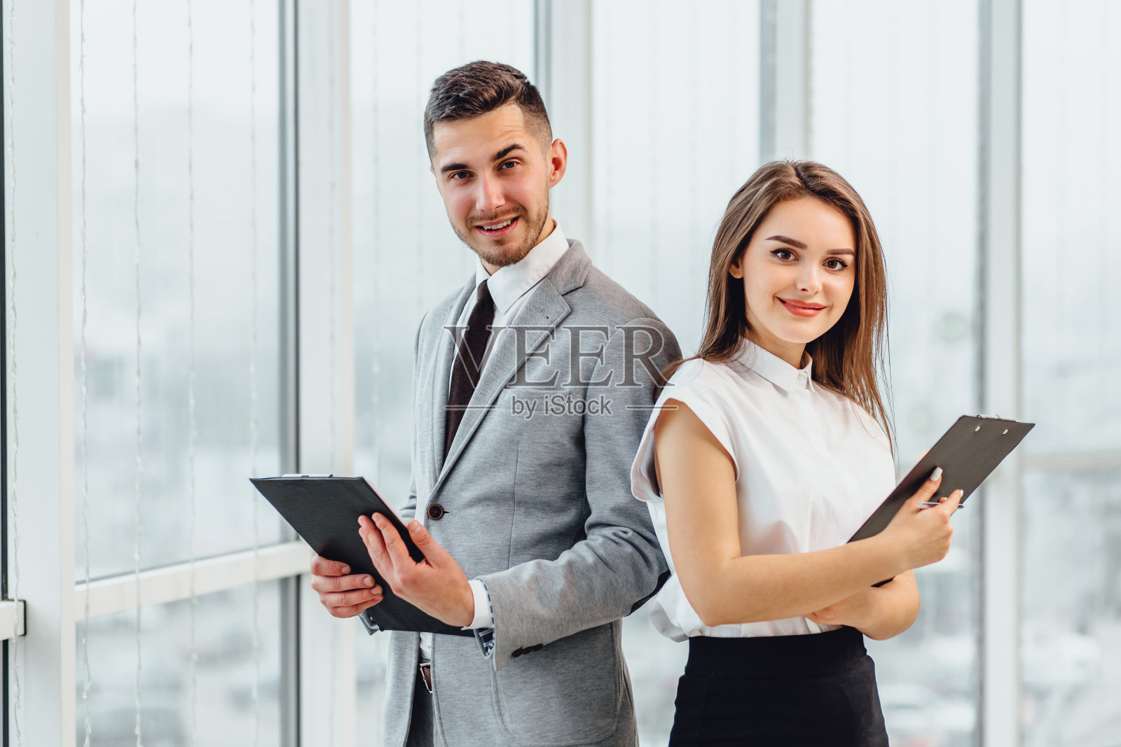 快乐的商业夫妇肩并肩站着，拿着剪贴板，看着镜头，微笑着。照片摄影图片