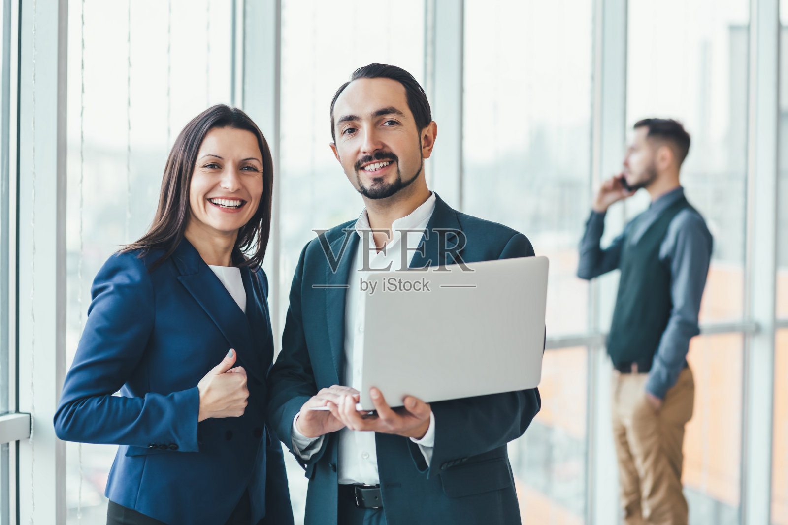 成功的男人和女人正在制定商业计划，在笔记本电脑中比较年度数据，而他们的同事正在模糊的背景下打电话。照片摄影图片