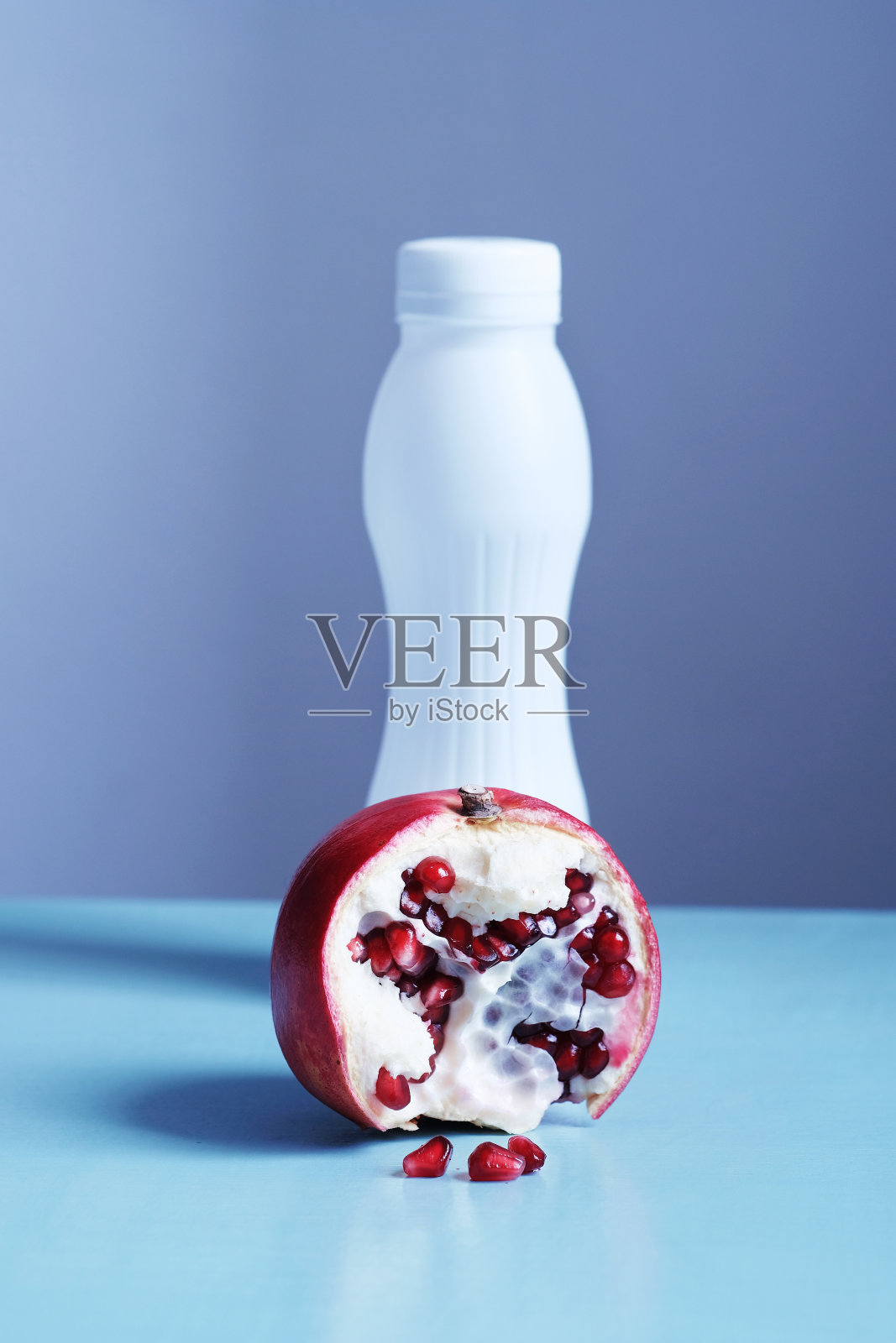 垂直拍摄的红色鲜亮的石榴和一个白色的酸奶瓶在背景上的蓝色桌子和灰色的背景。健康食品和饮料照片摄影图片