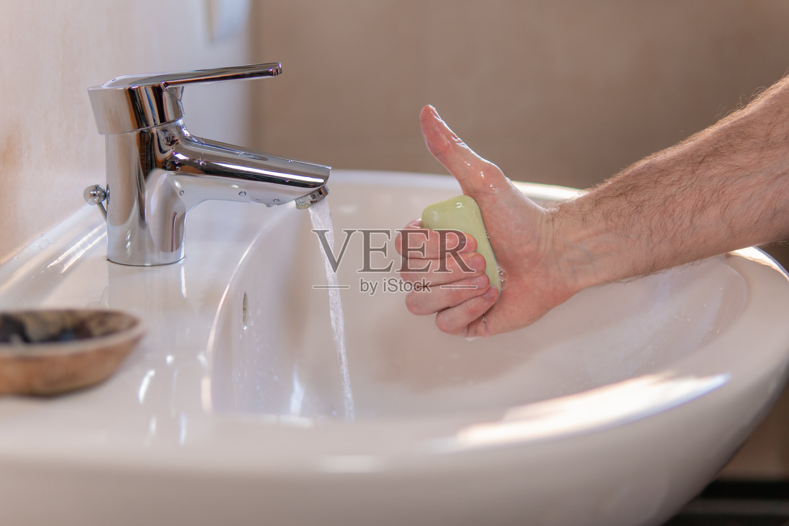 一个年轻白人男子的手拿着一块肥皂，并竖起大拇指。水龙头开着。个人卫生观念，节约用水，节约用水费用。照片摄影图片