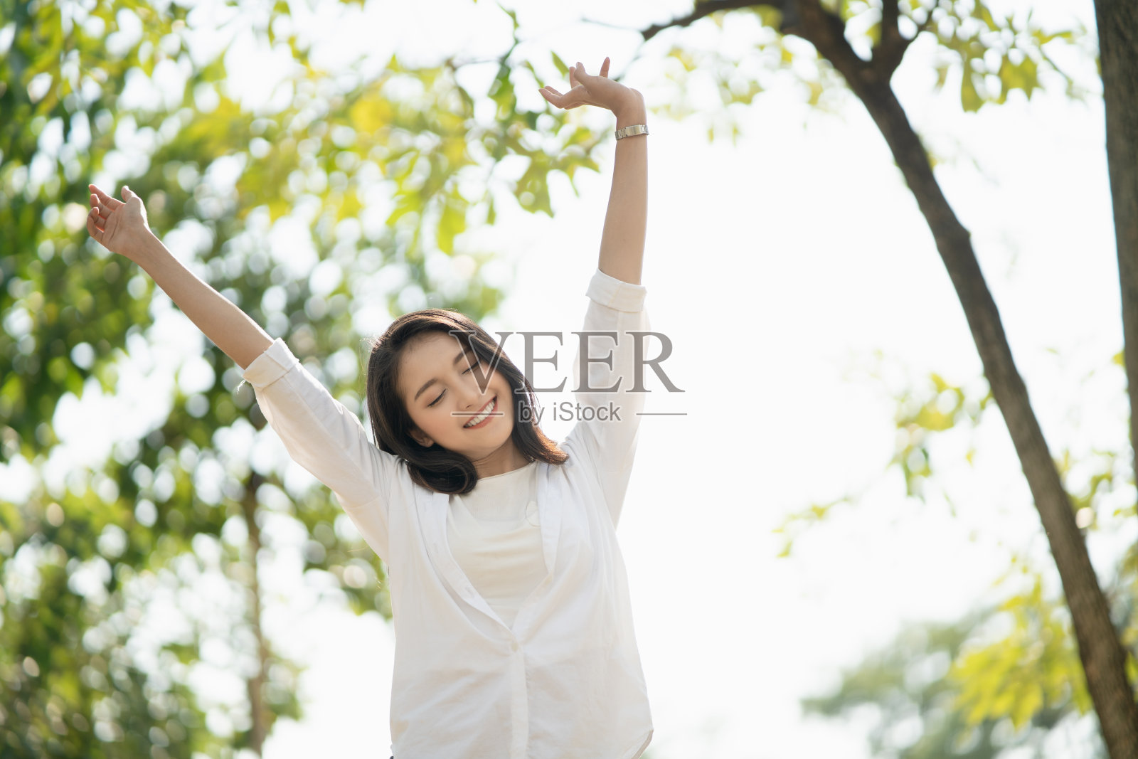 美丽的年轻亚洲女子举手微笑开心地感受悠闲放松在公园的自然阳光与复印空间，享受瞬间，遐想，悠闲自由的生活理念。照片摄影图片