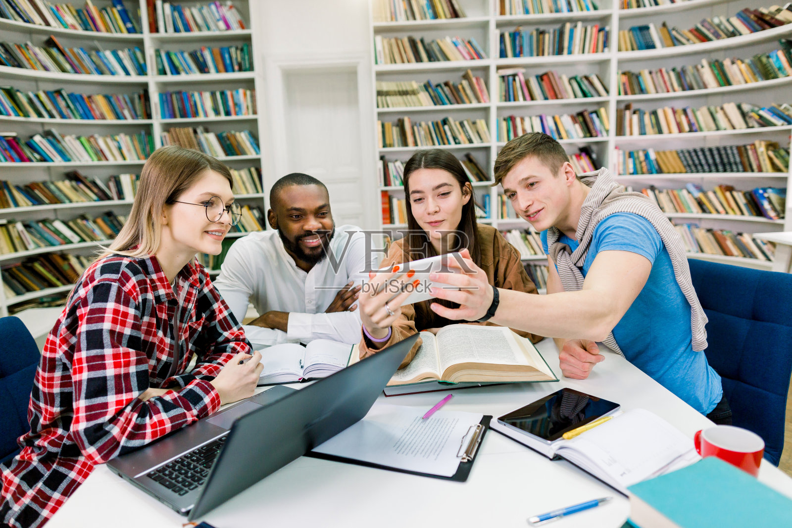 四个朋友，漂亮开朗快乐的混血儿学生，一边看智能手机上有趣的视频，一边在图书馆的书架上做小组学习的背景照片摄影图片