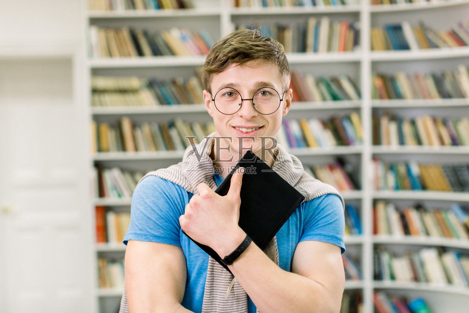 身穿蓝色t恤，戴着眼镜，手里拿着黑色的书，面带微笑的年轻学生站在图书馆现代阅览室的大书架前，看着相机照片摄影图片