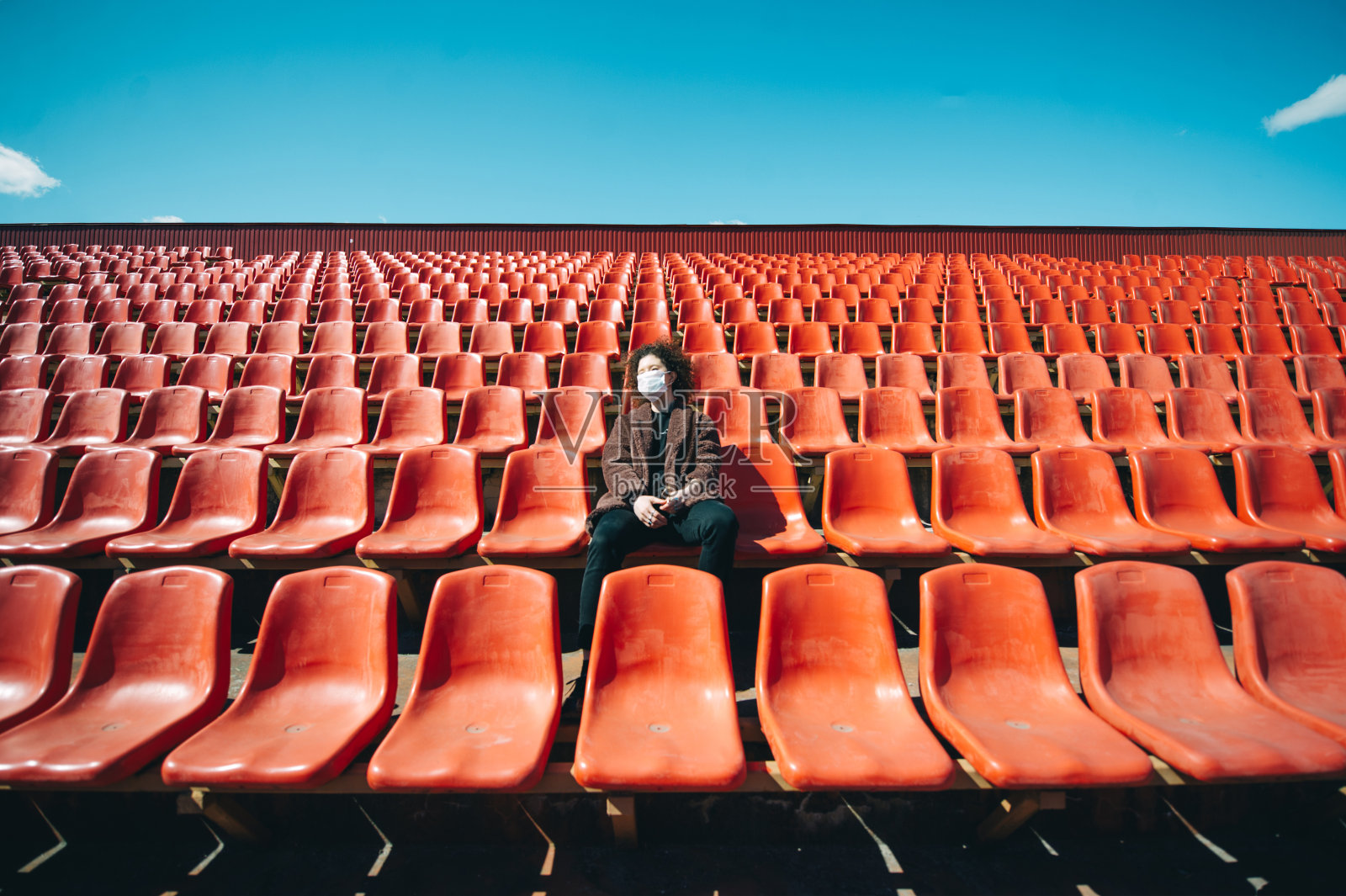 年轻的卷发女人戴着一次性医用口罩坐在空荡荡的城市体育场里。照片摄影图片