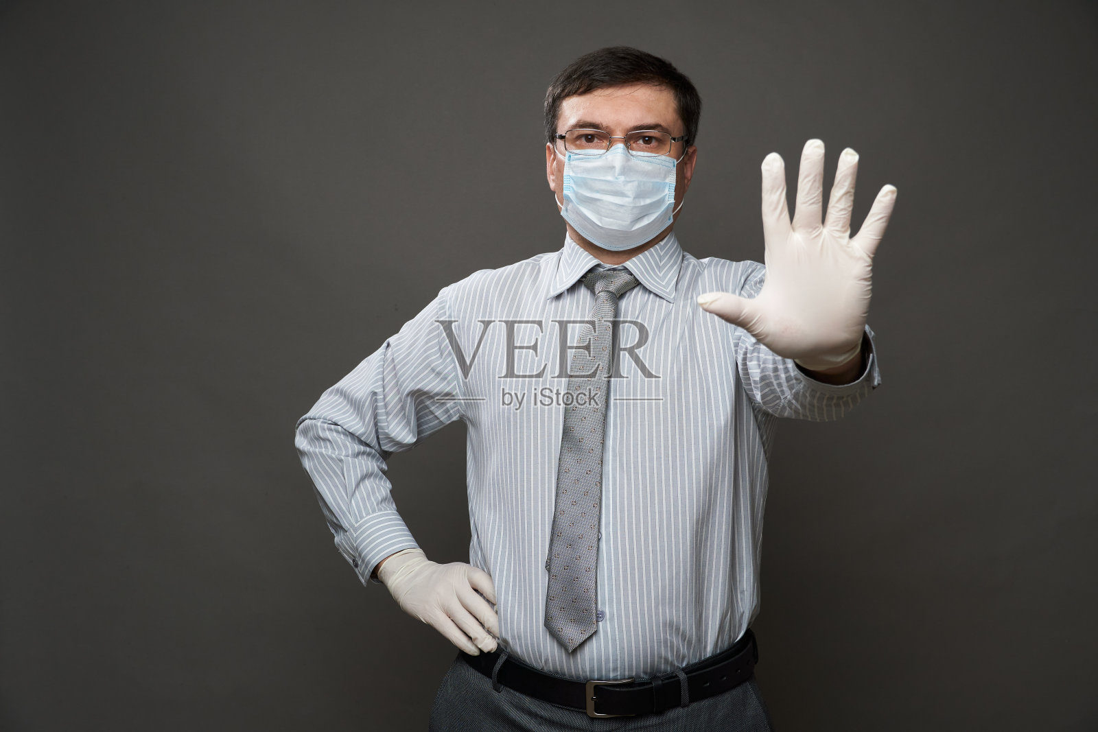 一名身着商人服装的男子用他的手掌、灰色背景、医用口罩和防护手套、眼镜、衬衫和领带——检疫和防病毒防护的概念照片摄影图片