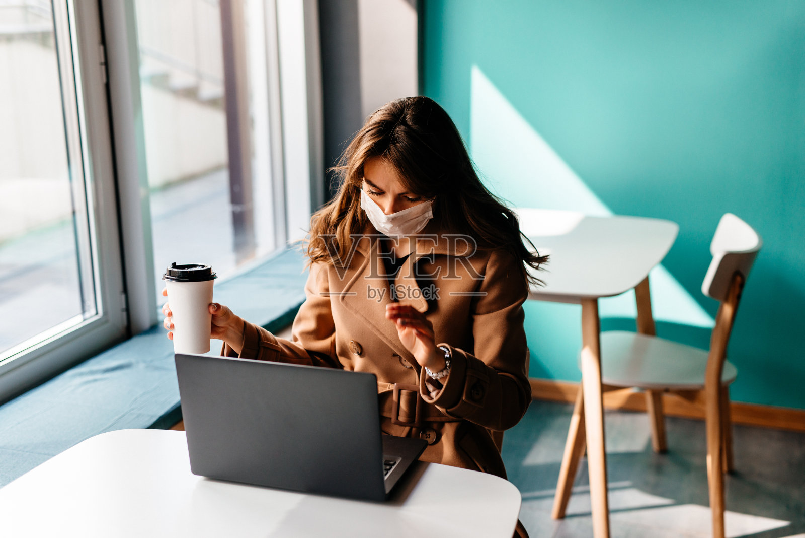 一名年轻女子戴着防护面具坐在咖啡馆的笔记本电脑前。一个女孩在一家咖啡店工作，她戴着黑色医用口罩和手套，手里拿着一杯咖啡照片摄影图片