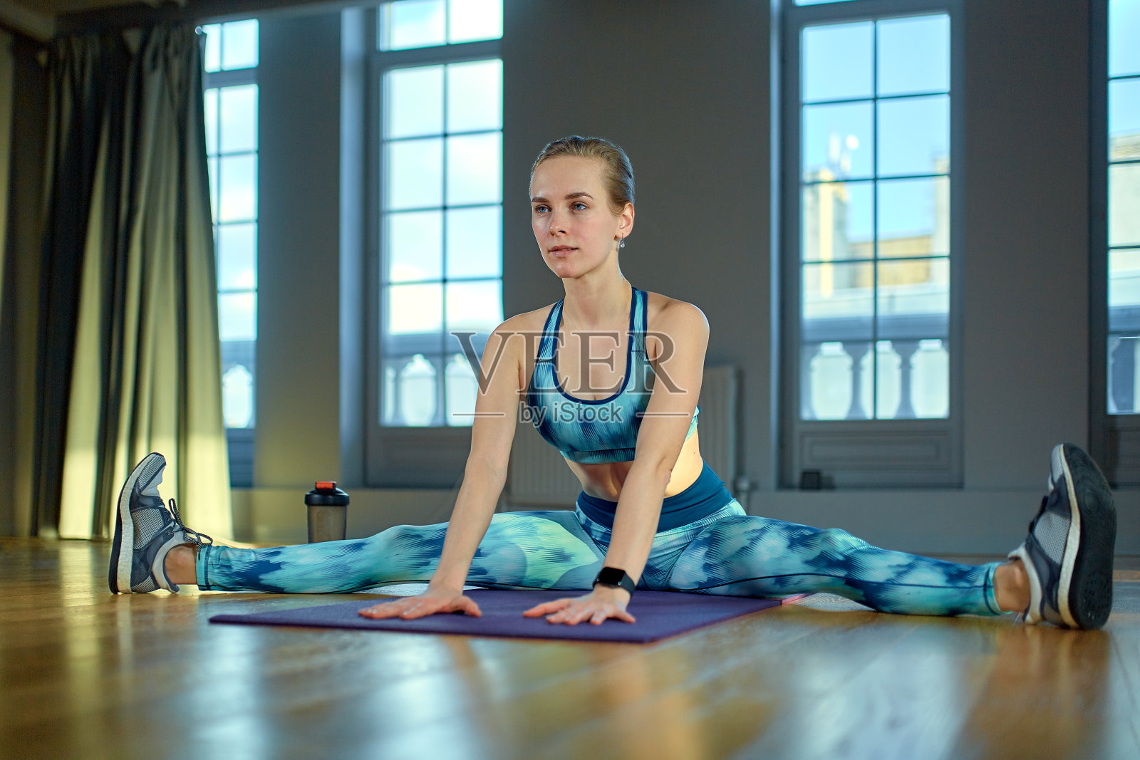保持灵活。年轻漂亮的女人穿着运动服，坐在健身房窗前的地板上做伸展运动照片摄影图片