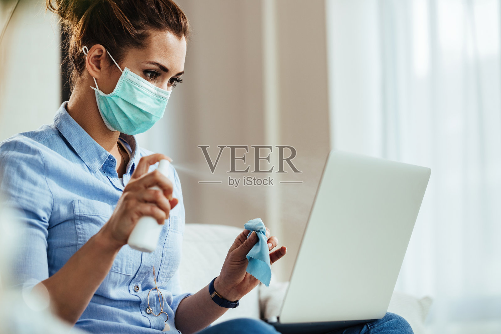 带着口罩在家给笔记本电脑消毒的女人。照片摄影图片