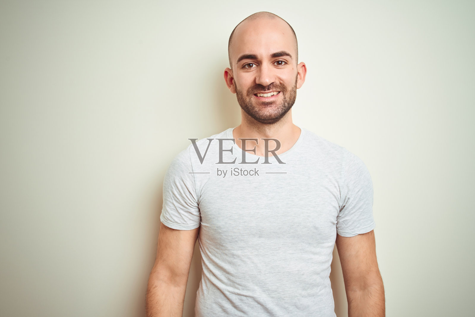 年轻的秃顶男子，留着胡子，穿着休闲的白色t恤，在孤立的背景下，脸上带着幸福和凉爽的微笑。幸运的人。照片摄影图片