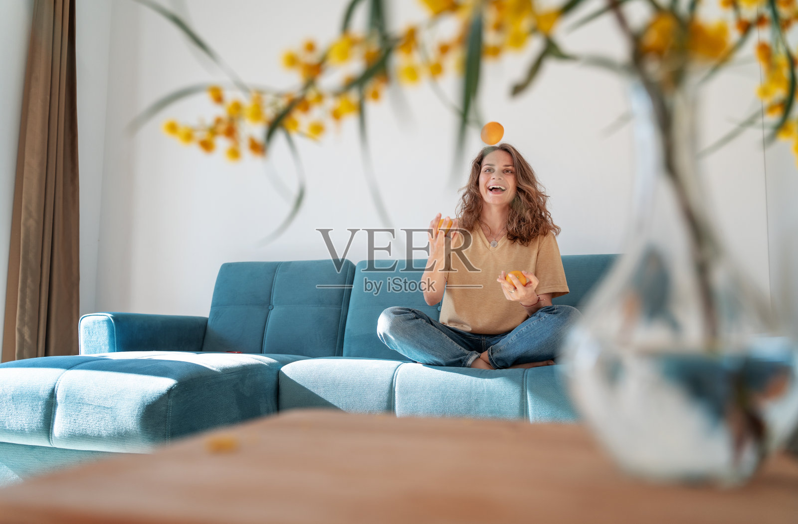 迷人的美丽的年轻女孩与卷曲的头发和一个幸福的微笑，而坐在她的客厅的蓝色沙发上的橘子，玩在家里的乐趣照片摄影图片