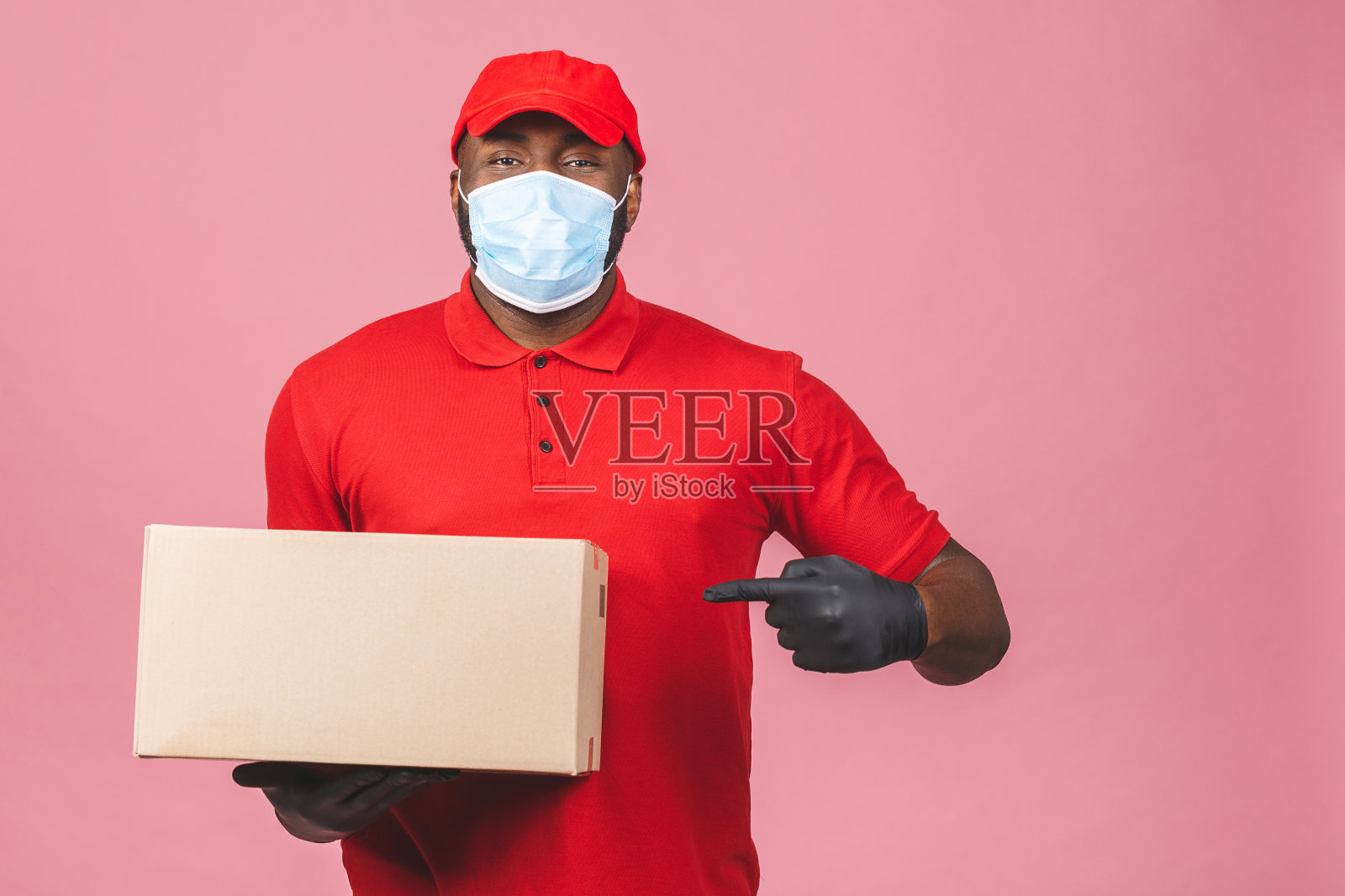 送货的非裔美国男子雇员在红色的帽子，空白的t恤制服，面罩，手套，拿空纸板箱隔离在粉红色的背景。服务隔离大流行性冠状病毒新型冠状病毒。照片摄影图片
