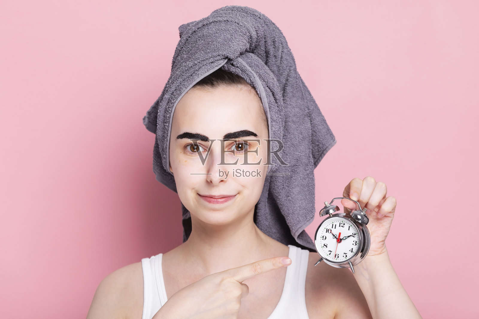 年轻女子头上裹着毛巾，自己画眉毛，手里拿着一个闹钟。居家和隔离的概念水疗日照片摄影图片