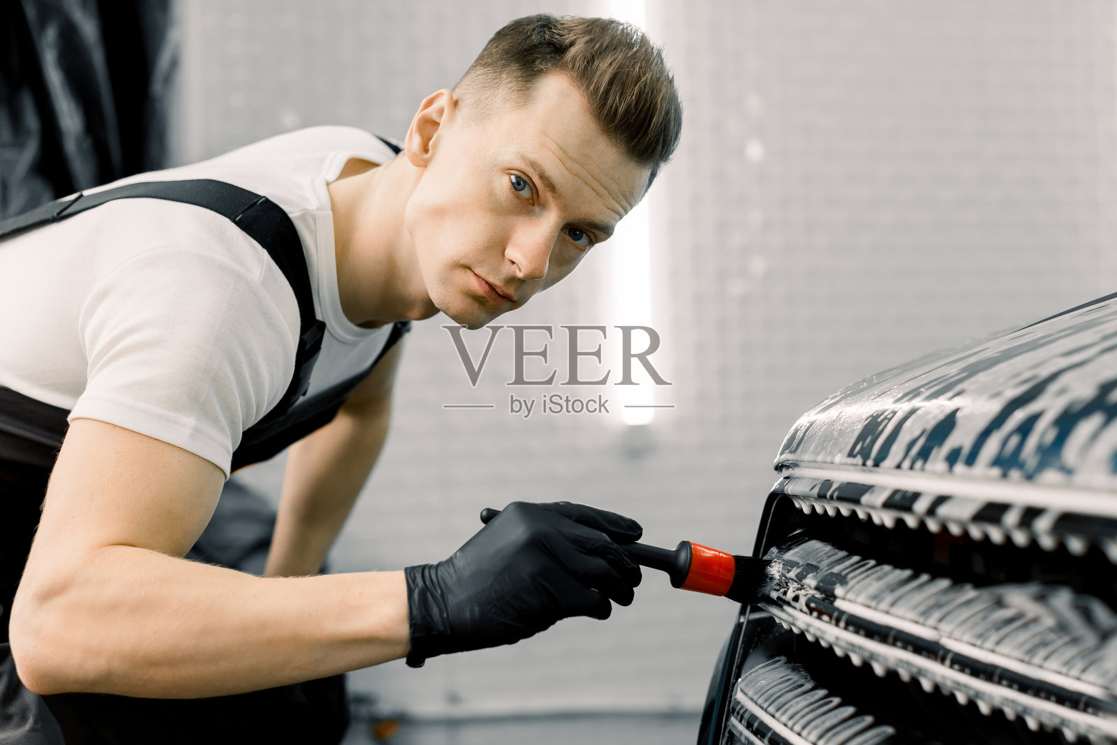 英俊的男性洗车工人看着摄像头，同时清洁汽车格栅与一个特殊的刷在汽车细节车间。汽车服务行业。照片摄影图片