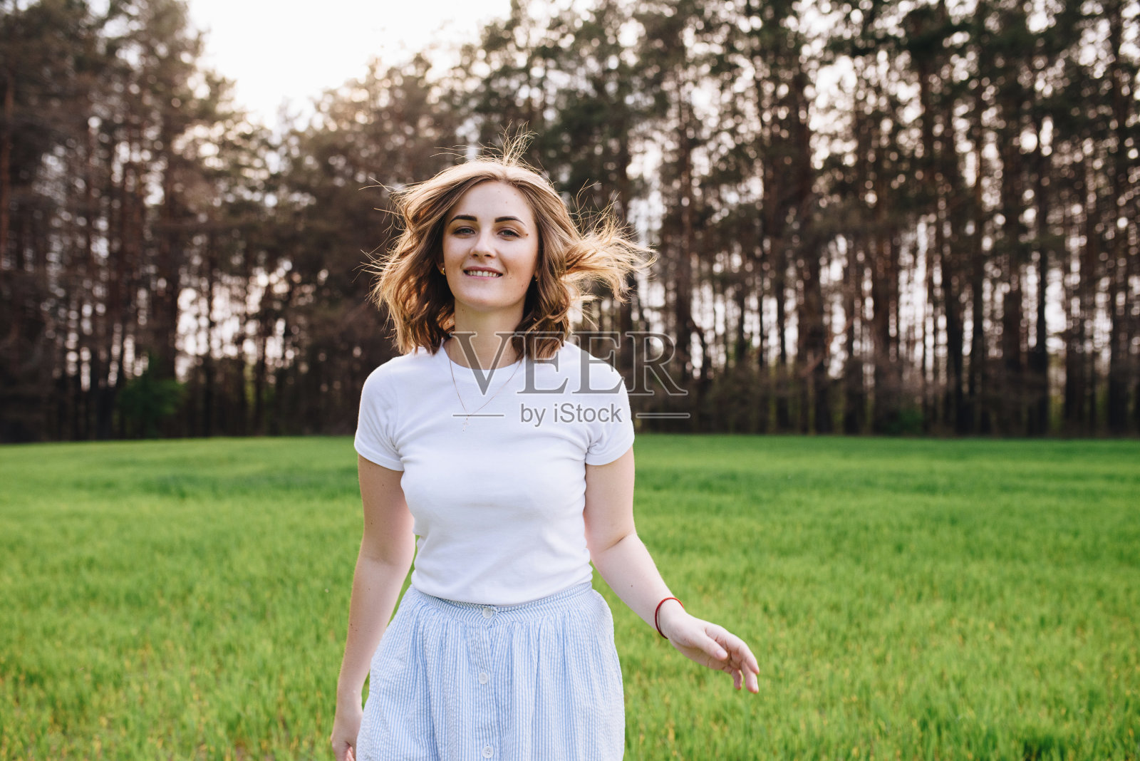 这个女孩金发棕发，穿着白色衬衫和蓝色中长裙。走在田野里，穿过绿色的草地。一个女孩的肖像。积极和微笑。照片摄影图片