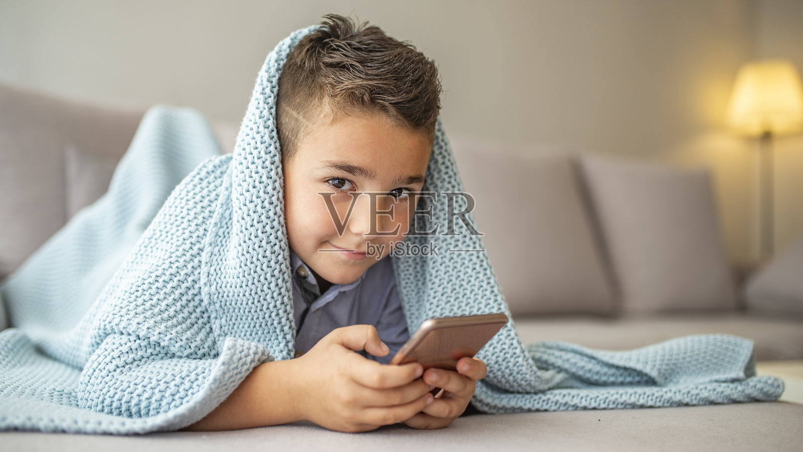 一个小男孩躺在沙发上，看着他的智能手机。照片摄影图片