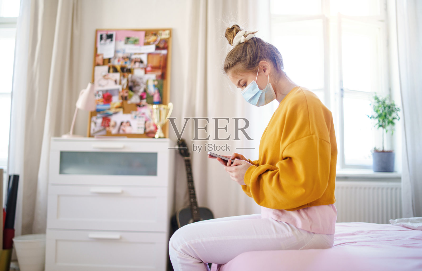 年轻女学生戴着口罩坐在床上，使用智能手机。照片摄影图片