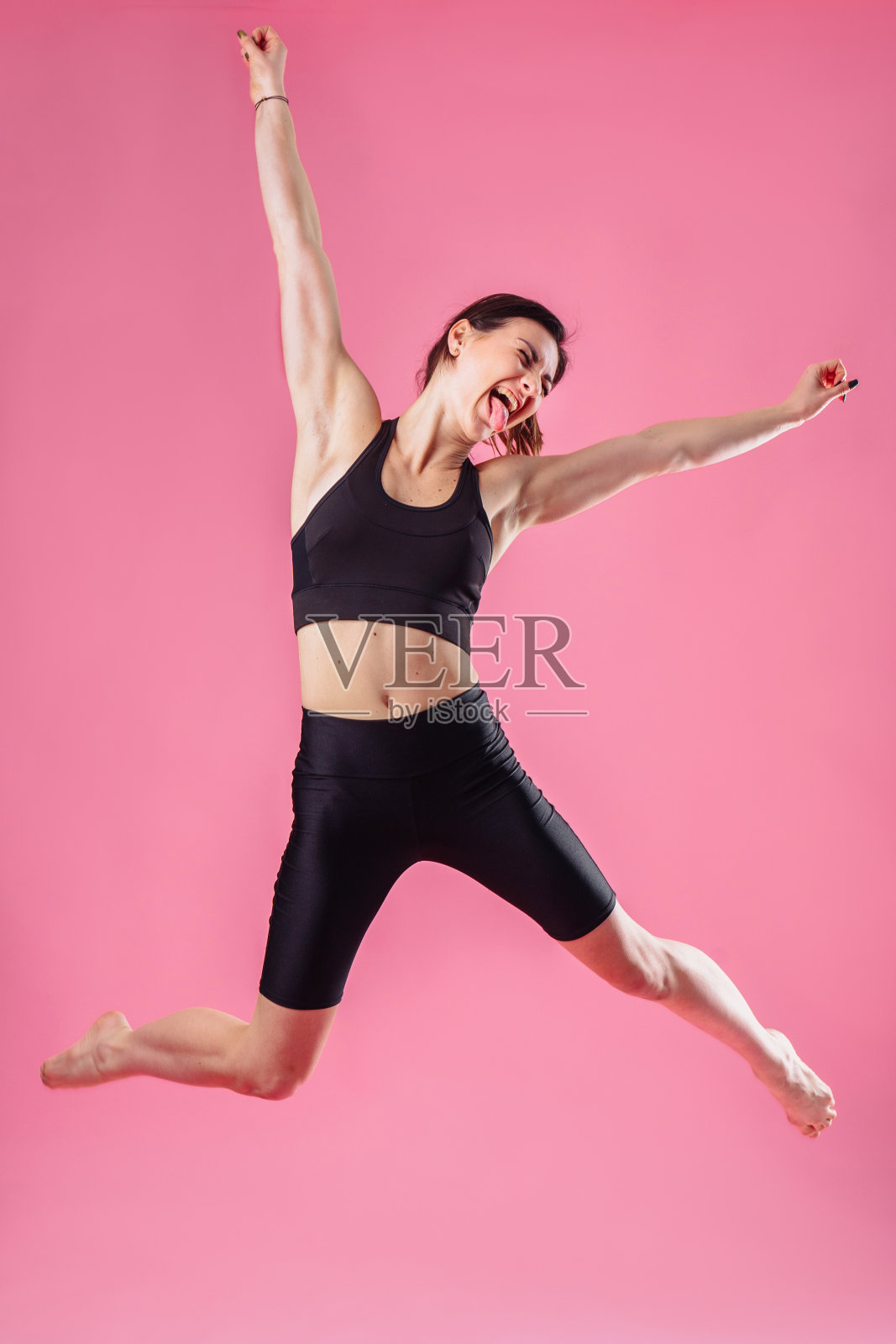 肌肉美丽的健身女孩在一个黑色的顶部孤立在粉红色的背景。快乐地跳起来，手和腿分开，尖叫。为她的高成绩感到高兴和高兴。照片摄影图片