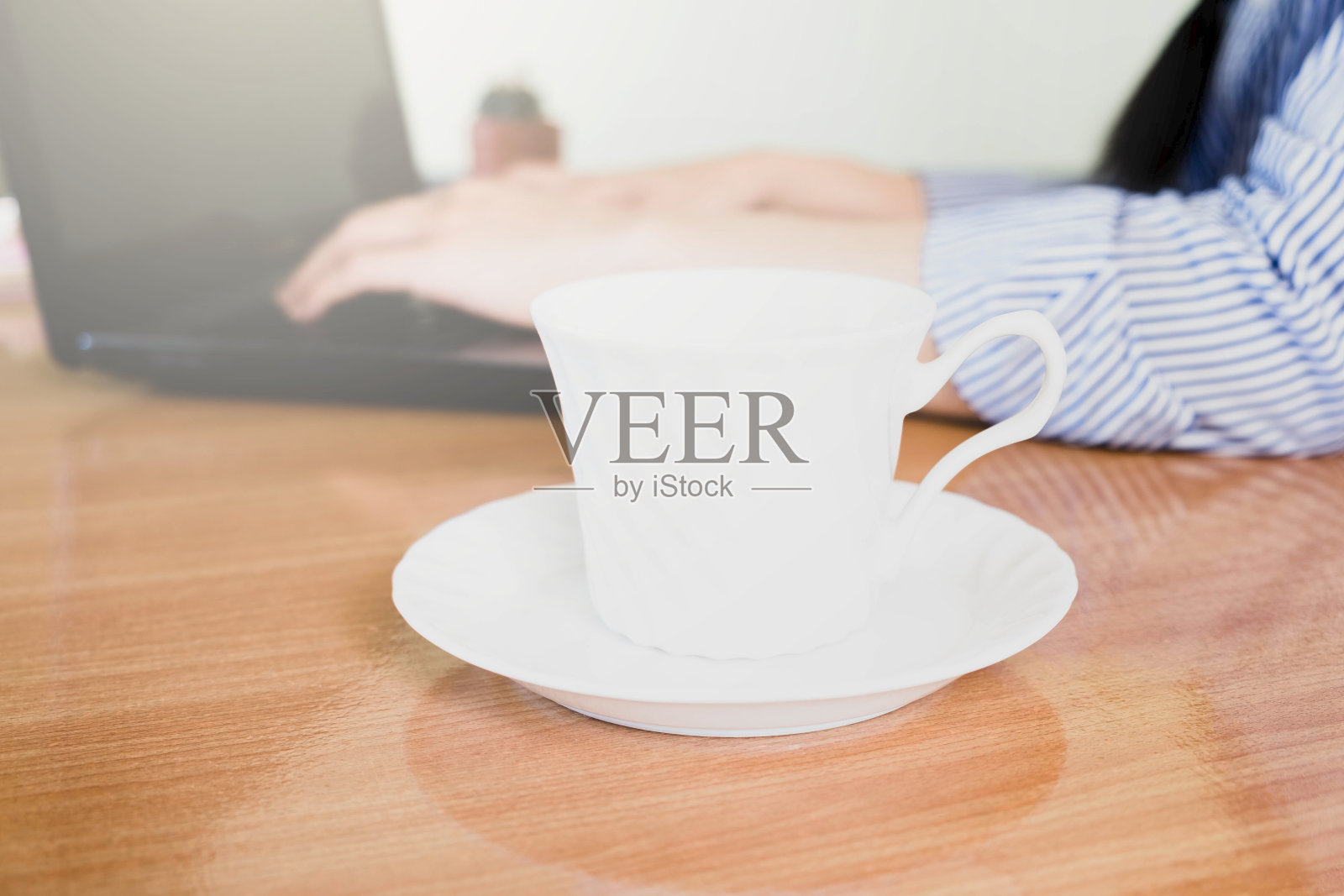 选择性聚焦白色的一杯咖啡，模糊女人的手穿着蓝色条纹衬衫在木桌上敲击键盘笔记本电脑，在线工作的生活方式，在家喝咖啡放松，在家工作的概念照片摄影图片