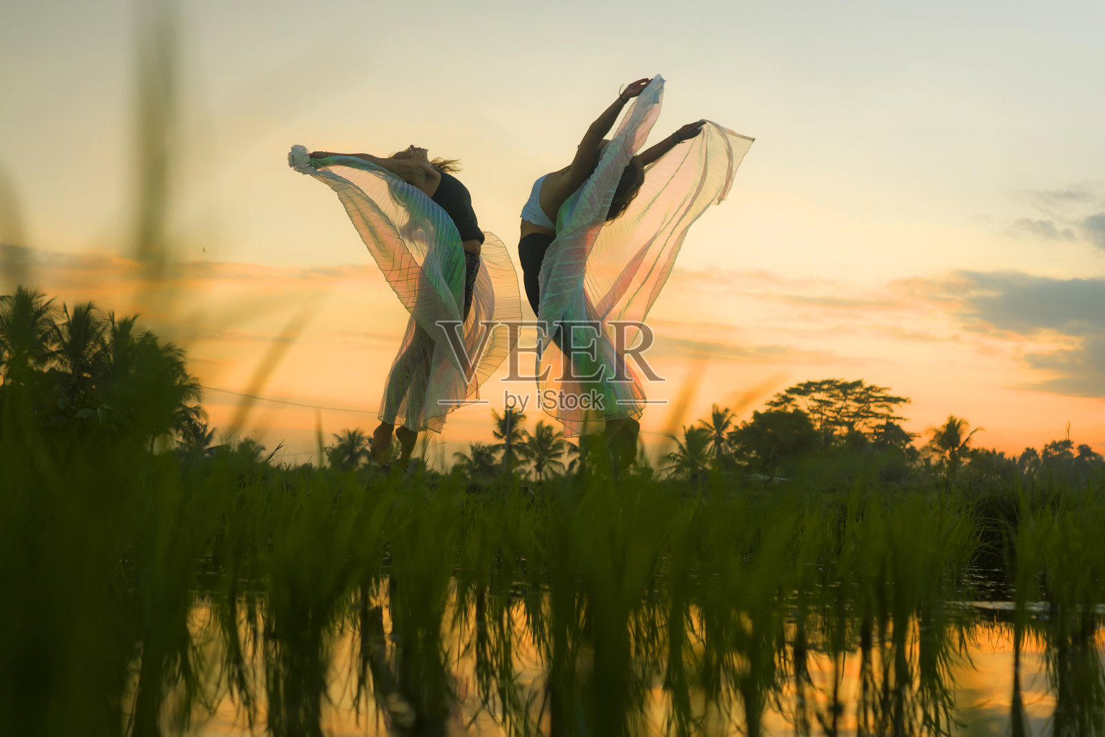 户外日落acroyoga锻炼-一对年轻快乐和健康的妇女练习跨越瑜伽与翅膀在美丽的稻田享受自然做杂技姿势，有乐趣照片摄影图片