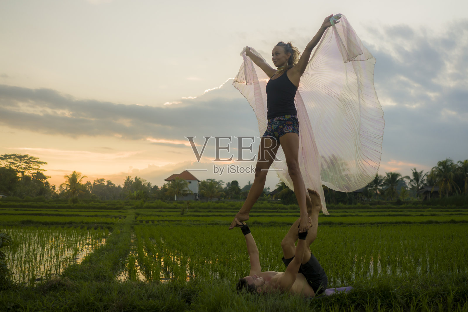 户外日落acroyoga锻炼-年轻快乐和健康的夫妇练习跨越瑜伽练习在美丽的稻田享受自然和健康的生活方式做杂技姿势照片摄影图片