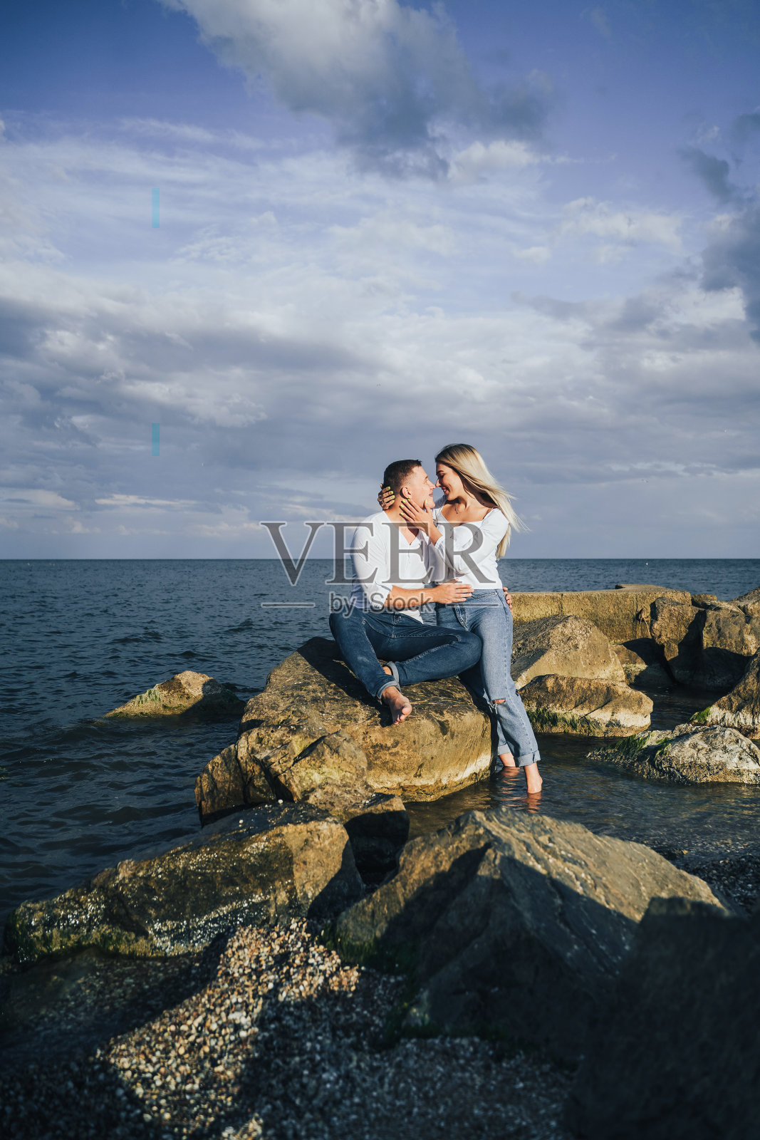 长针。一对迷人的夫妇穿着鲜艳的衣服站在海上的石头上，对抗着蓝色的多云的天空。被柔和的夕阳照耀着。期待，拥抱，亲吻，享受美丽的夜晚照片摄影图片