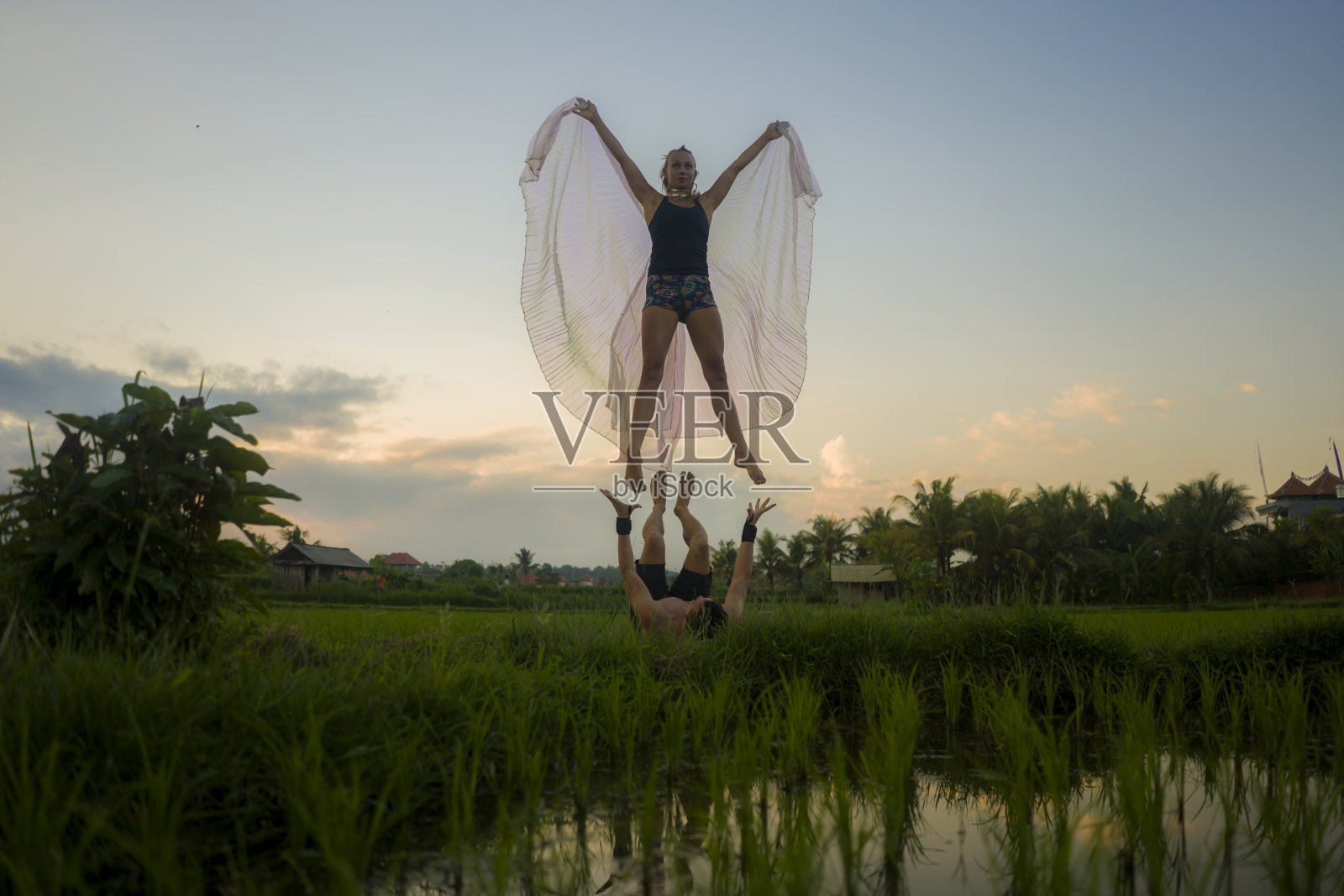 户外日落acroyoga锻炼-年轻快乐和适合夫妇练习跨越瑜伽练习在美丽的稻田享受自然做杂技姿势的女人在自由的概念翅膀照片摄影图片