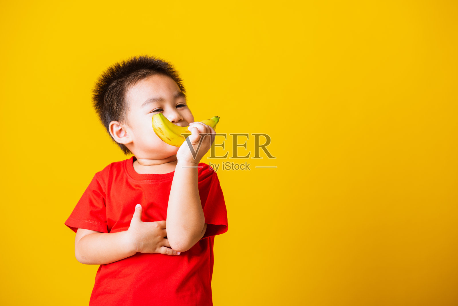 快乐肖像亚洲小孩或小孩可爱的小男孩迷人的微笑穿着红色t恤玩抱着香蕉水果，工作室拍摄孤立在黄色的背景照片摄影图片
