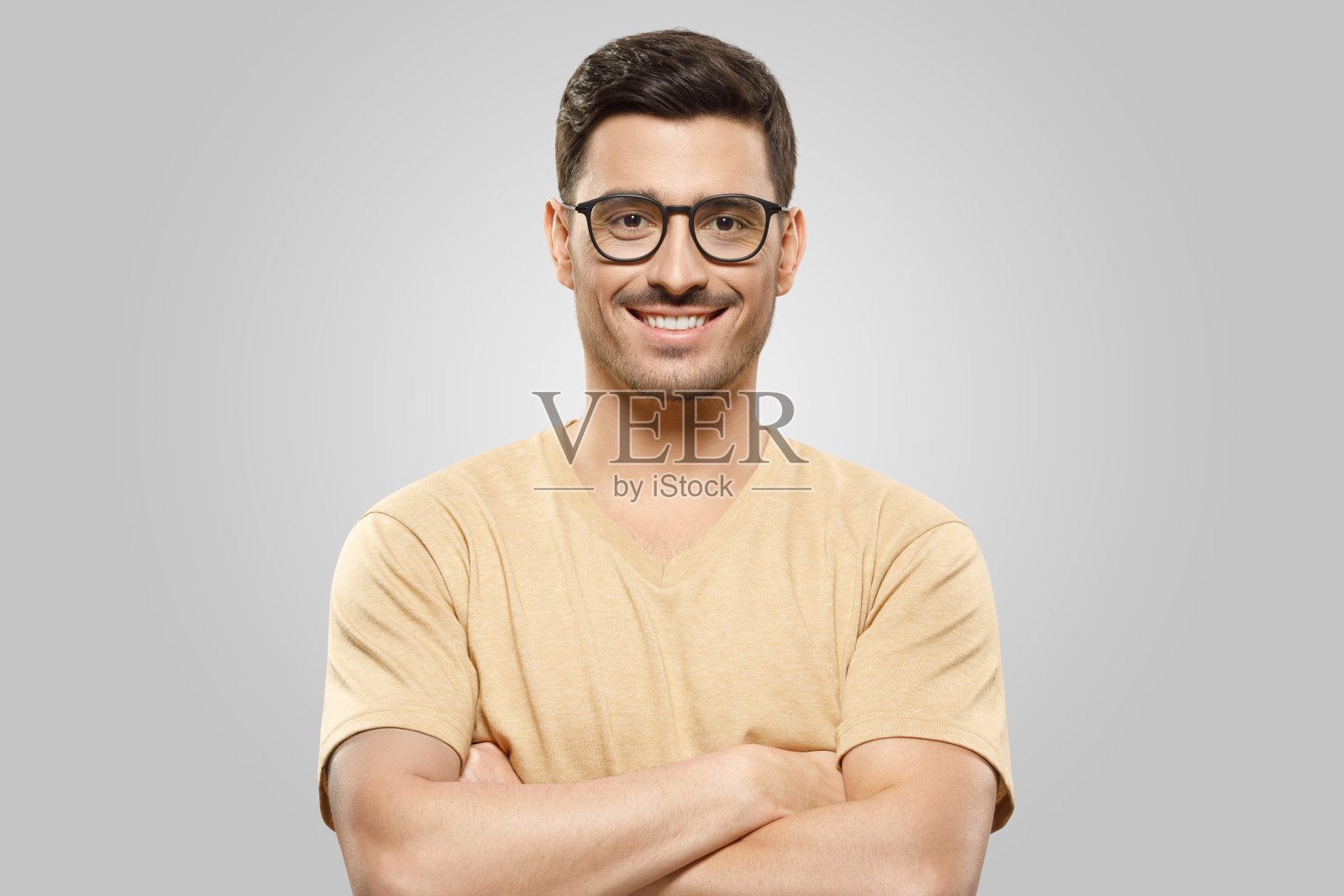 一个自信的年轻人双手交叉站着，戴着眼镜，穿着休闲的米色t恤，孤立在灰色的背景照片摄影图片