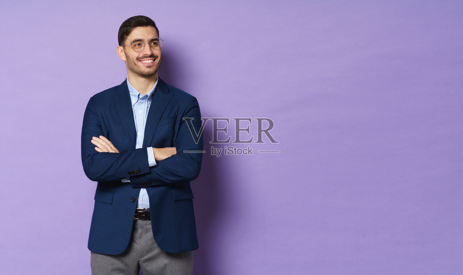 横向拍摄的年轻的商业男子站在夹克和眼镜，双臂交叉，看着一边，积极的微笑，紫色的背景，孤立的copyspace在右边照片摄影图片