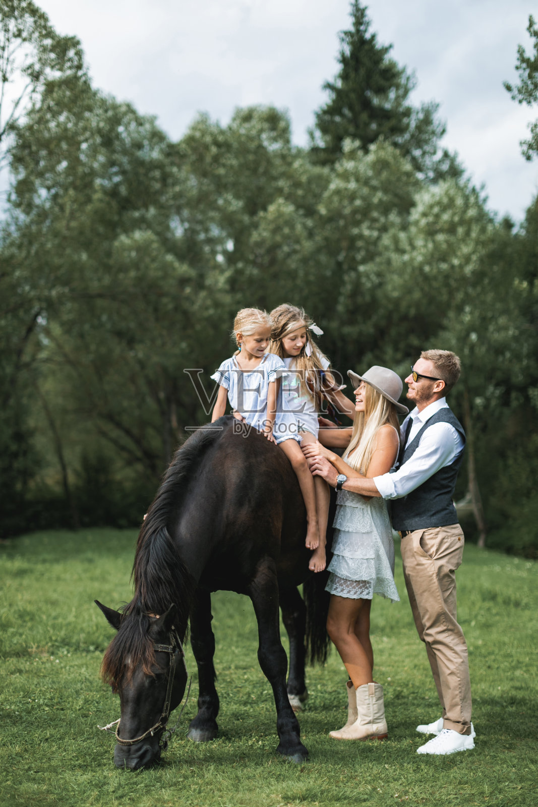 年轻时尚的家庭穿着牛仔装，父亲，母亲教他们的两个小女儿骑马。家骑马在大自然的乡间照片摄影图片