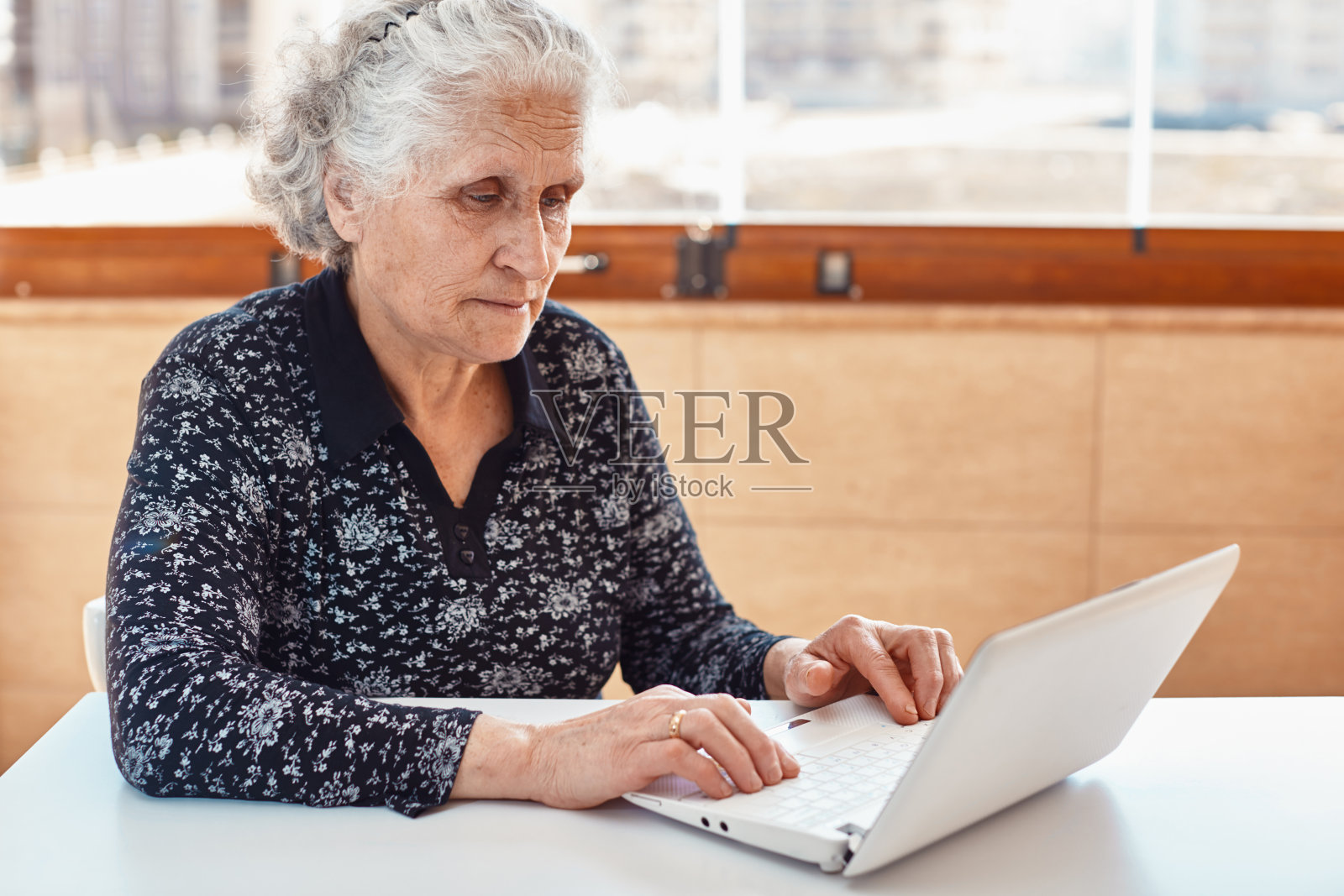 美丽的白发老人使用笔记本电脑在桌子上，城市的天际线为背景。照片摄影图片