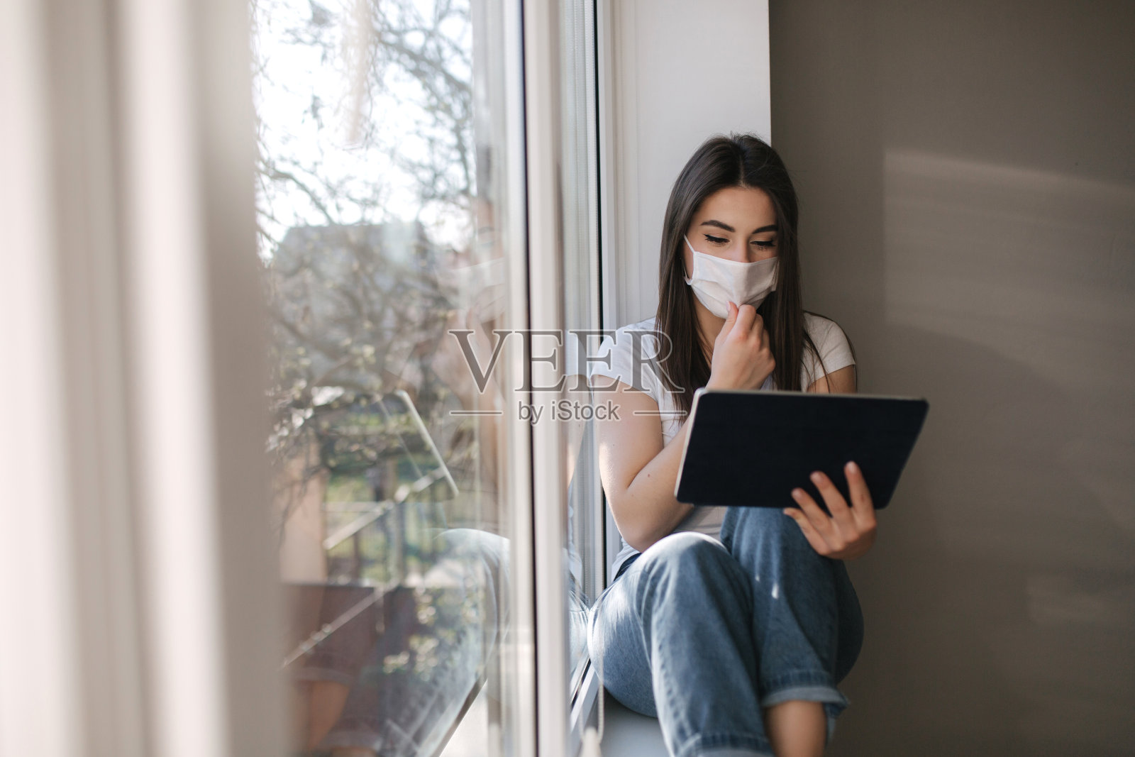 迷人的年轻女子戴着白色医用面具坐在窗台上，用平板电脑。女性在隔离期间在家休息照片摄影图片