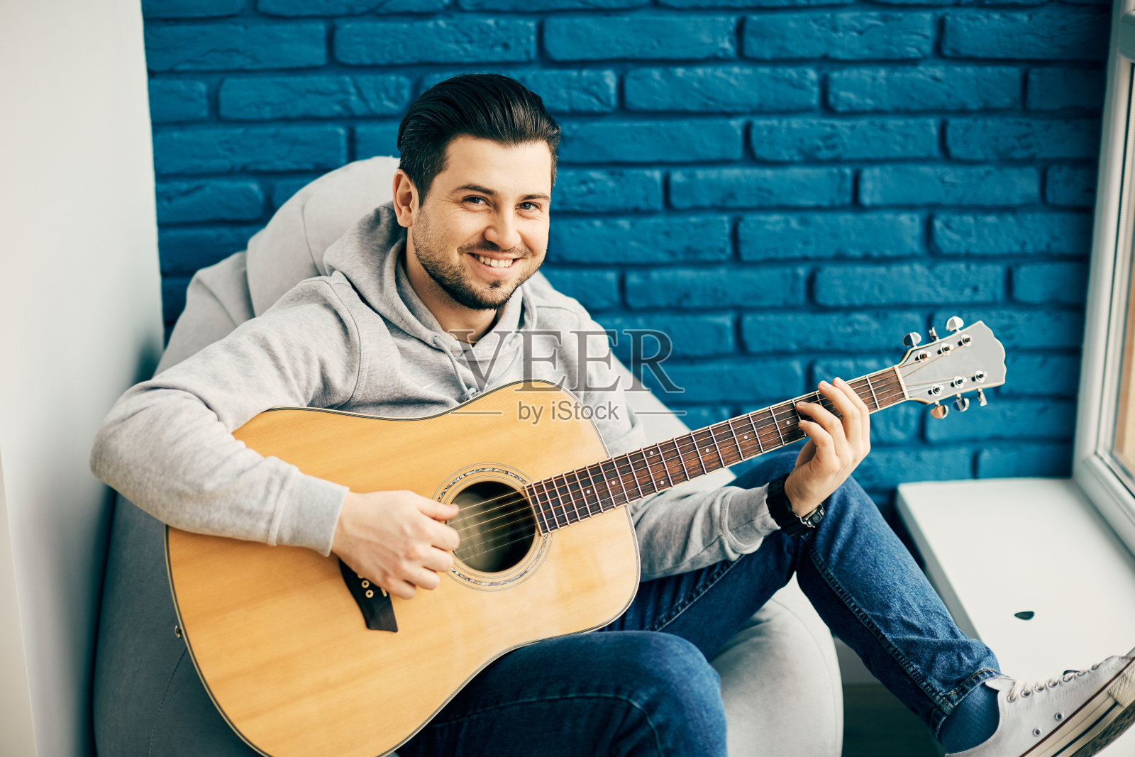 身穿灰色毛衣的英俊男子拿着吉他，背景是蓝色砖墙，对着镜头微笑照片摄影图片